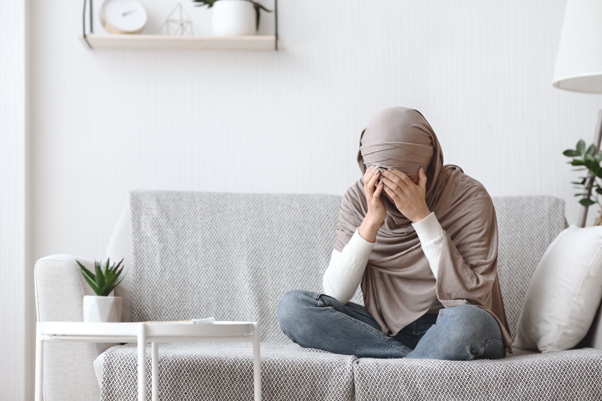 Upset muslim woman in headscarf crying on couch at home